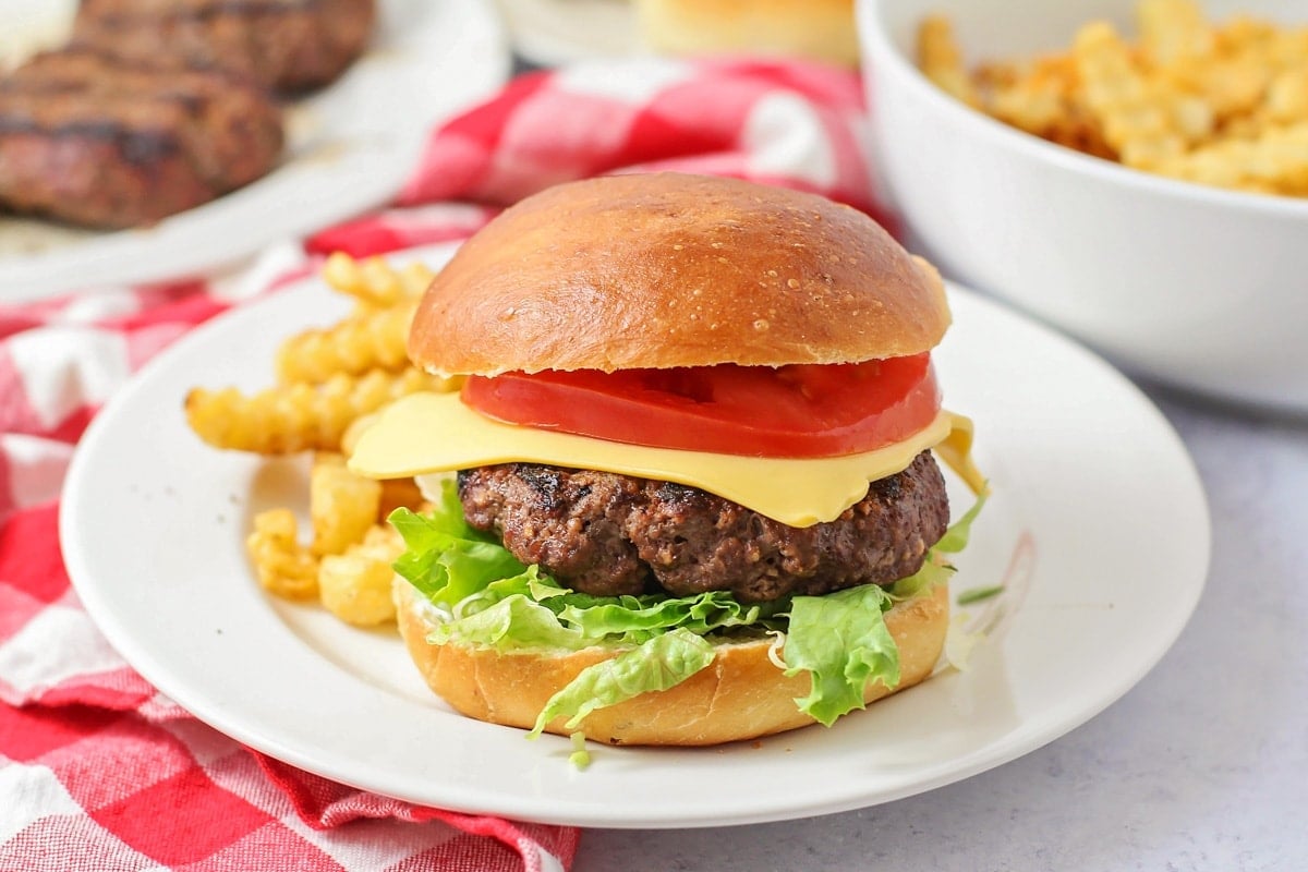 A cheeseburger with lettuce and tomato on a bun next to french fries on a white plate with a red and white checked tablecloth, more burgers and more french fries in the background.