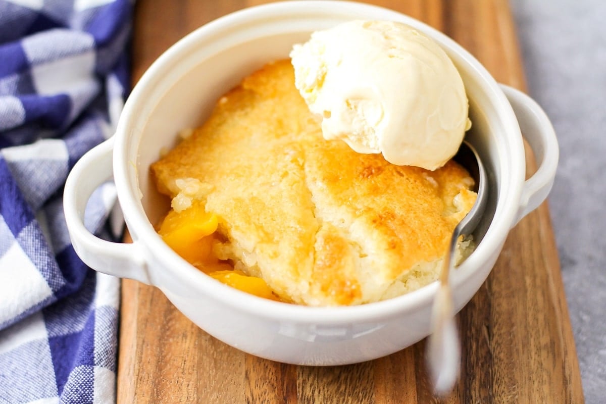 Peach Cobbler topped with a scoop of vanilla ice cream in a small white bowl with a silver spoon resting in the bowl. 