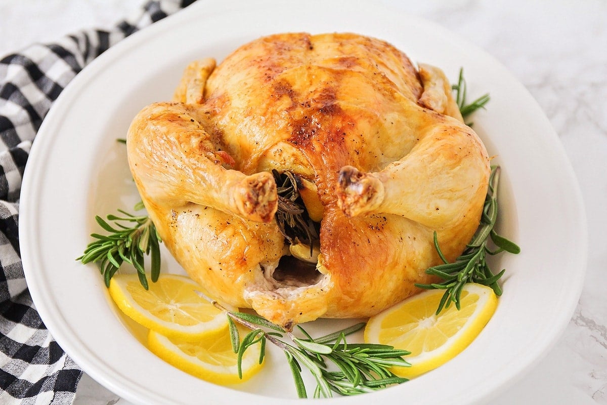 Roast chicken garnished with lemon slices and rosemary sprigs on a white platter. 