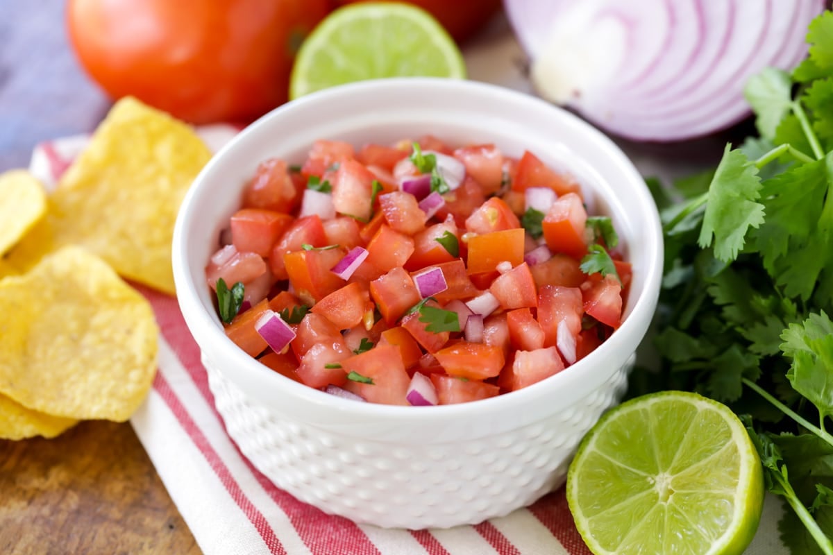 Pico de Gallo in a small white bowl surrounded by tortilla chips and fresh produce.