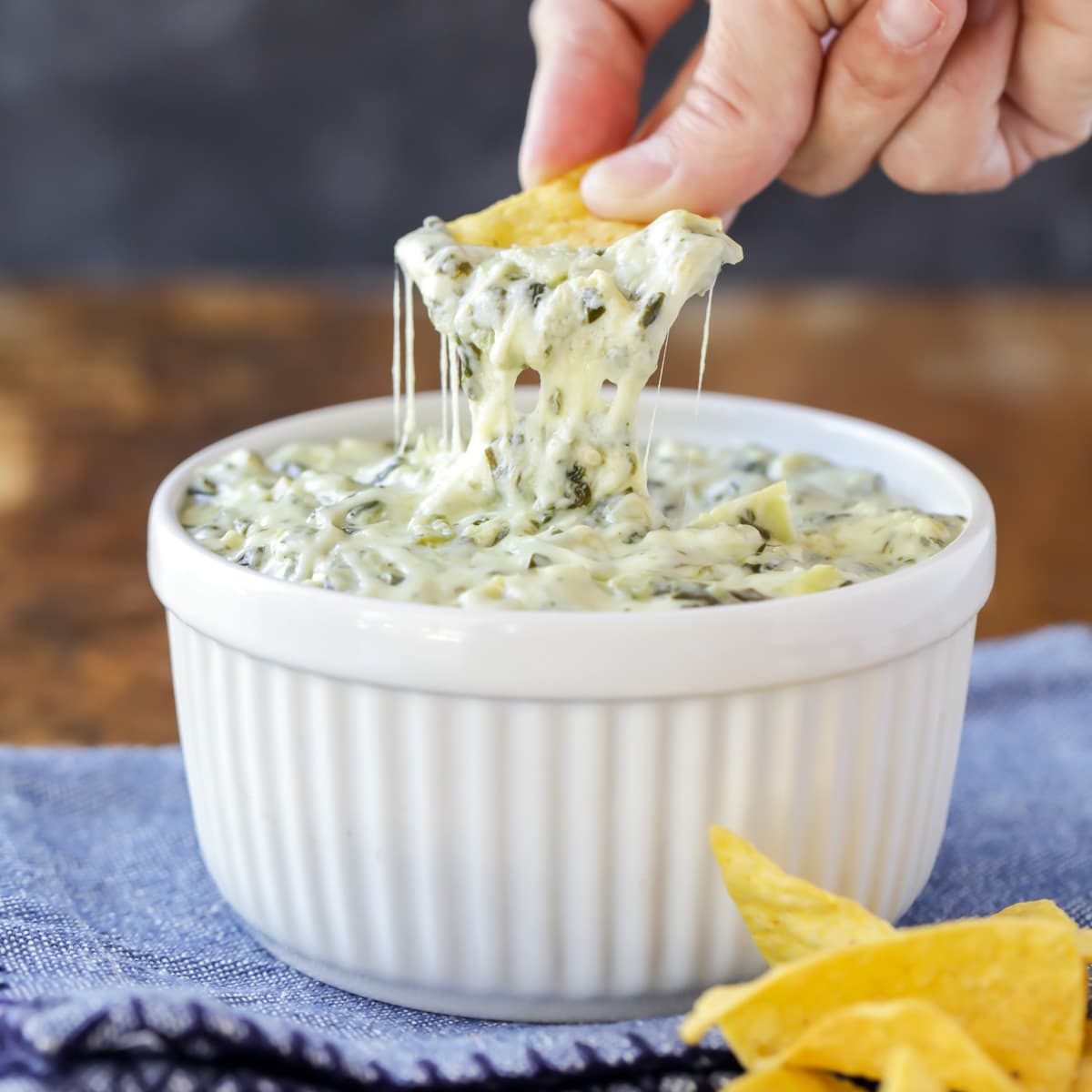 A hand dipping a chip into a bowl of spinach artichoke dip.