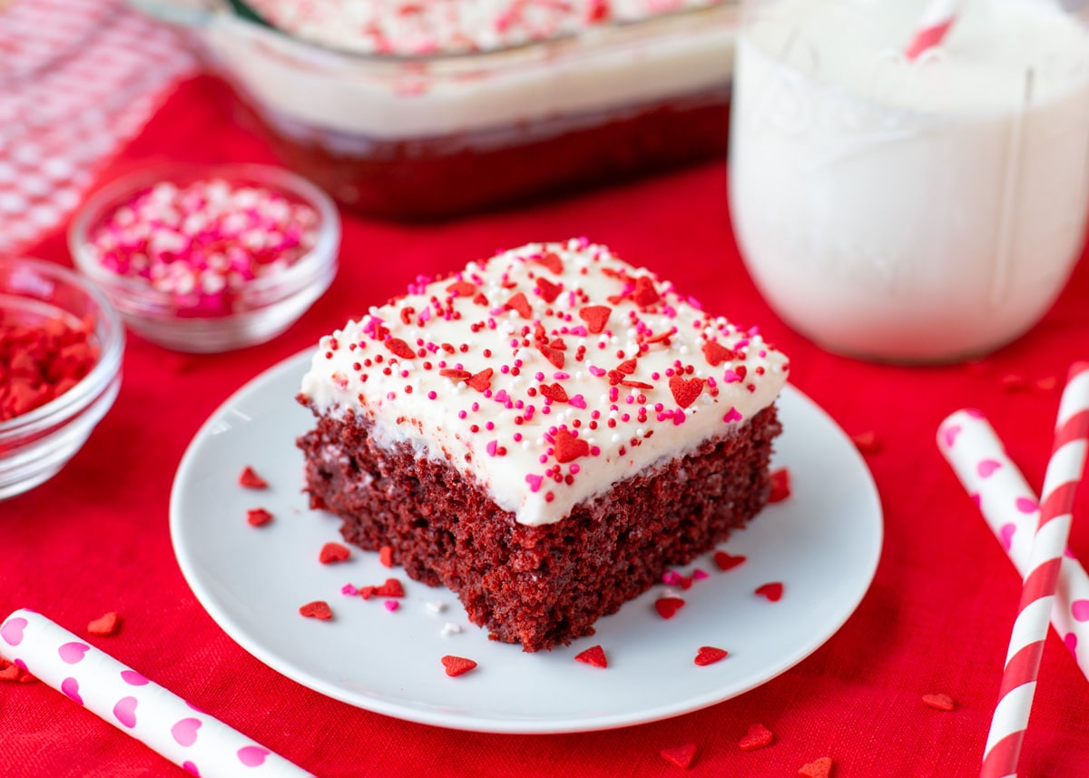 Valentines Dinner Ideas - red velvet poke cake with white frosting and pink and red sprinkles on a white plate. 