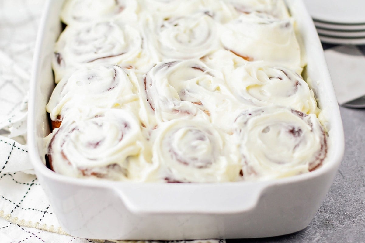 A white baking dish full of cinnamon rolls topped with cream cheese frosting.