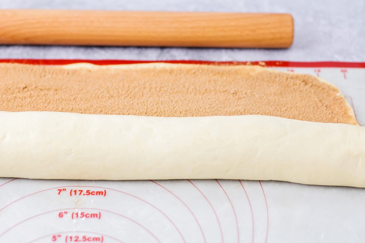 Cinnamon roll dough filled with a cinnamon brown sugar mixture being rolled up on a plastic mat with a wooden rolling pin in the background. 