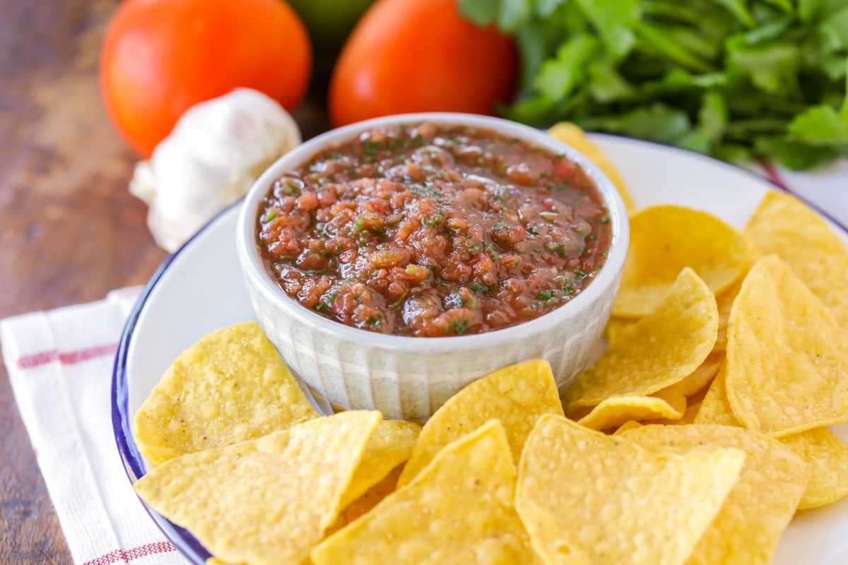 A white bowl filled with homemade salsa surrounded by tortilla chips on a white plate with blue around the rim.