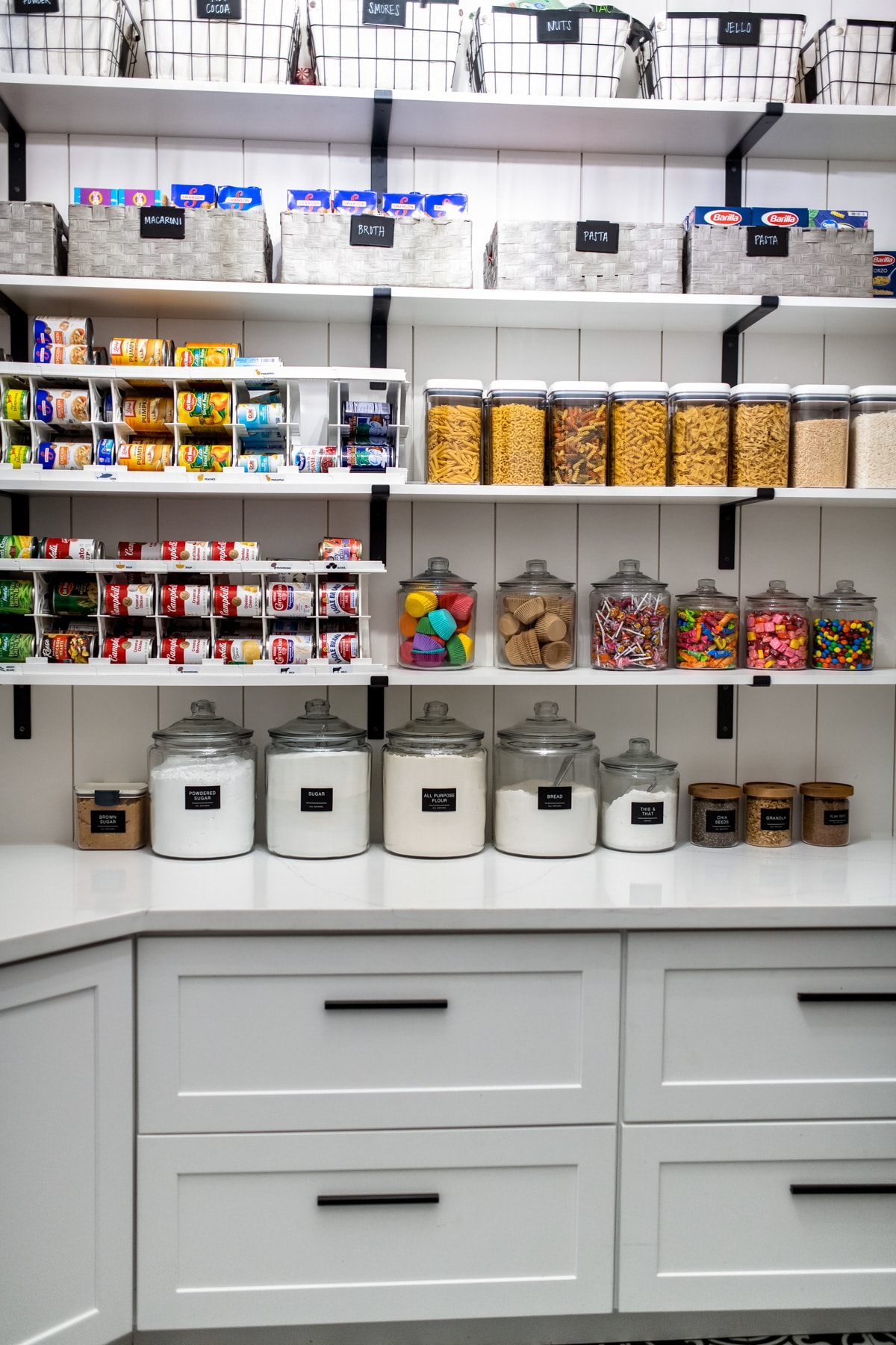 An organized pantry with labeled baskets, can organizers, food storage containers, and labeled jars lining the shelves.