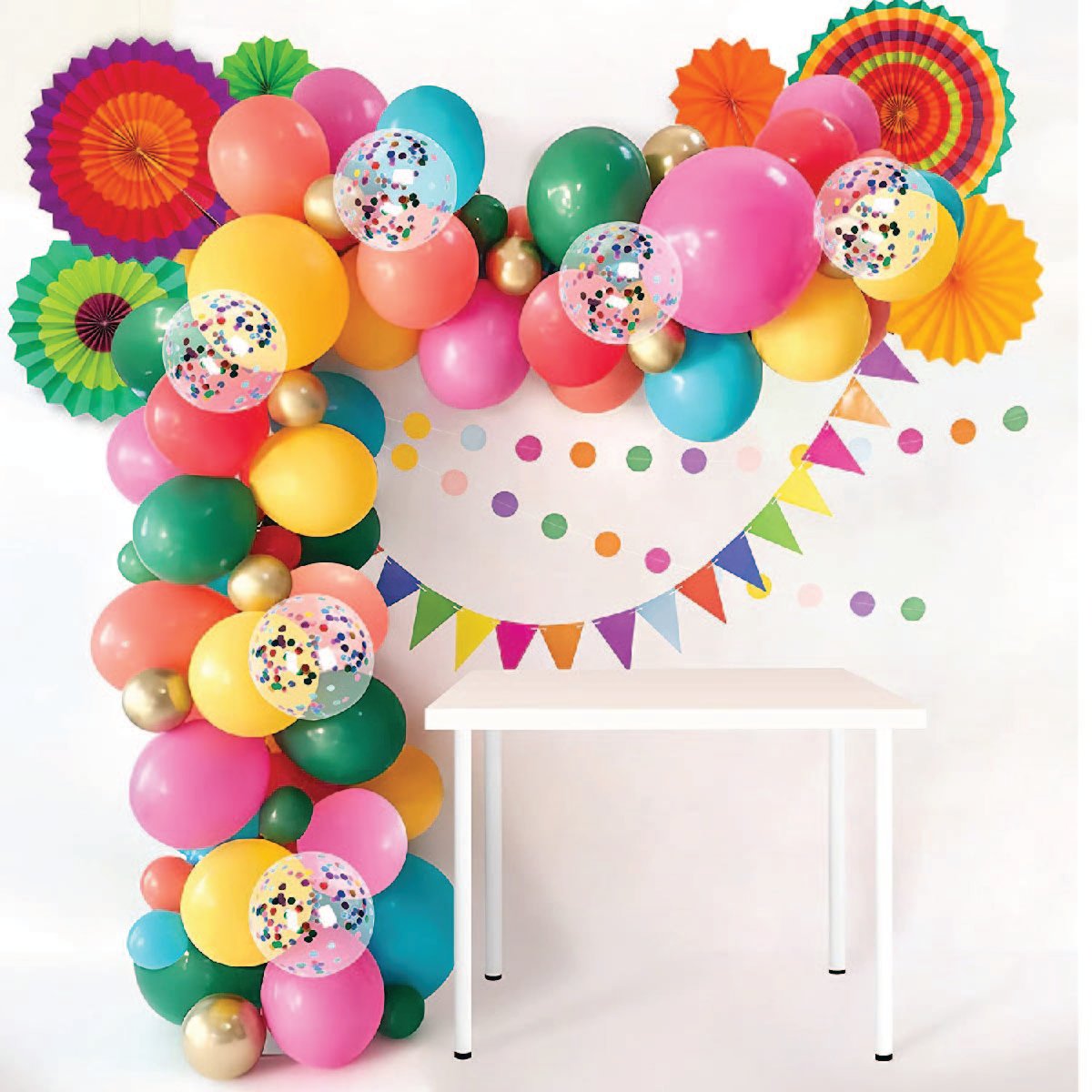 A strand of several different colored balloons with paper fans and banners against a white wall and above a small white table.