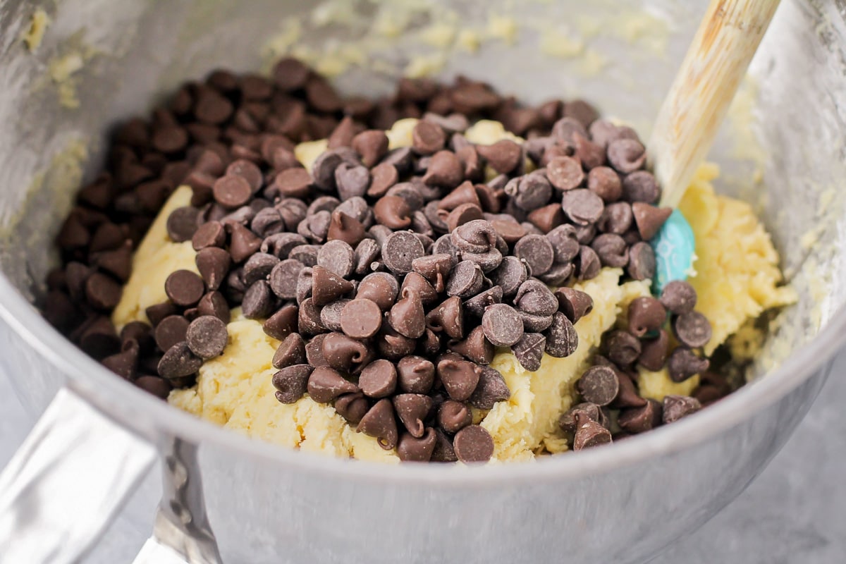 Adding chocolate chips to cookie dough batter in a metal bowl.