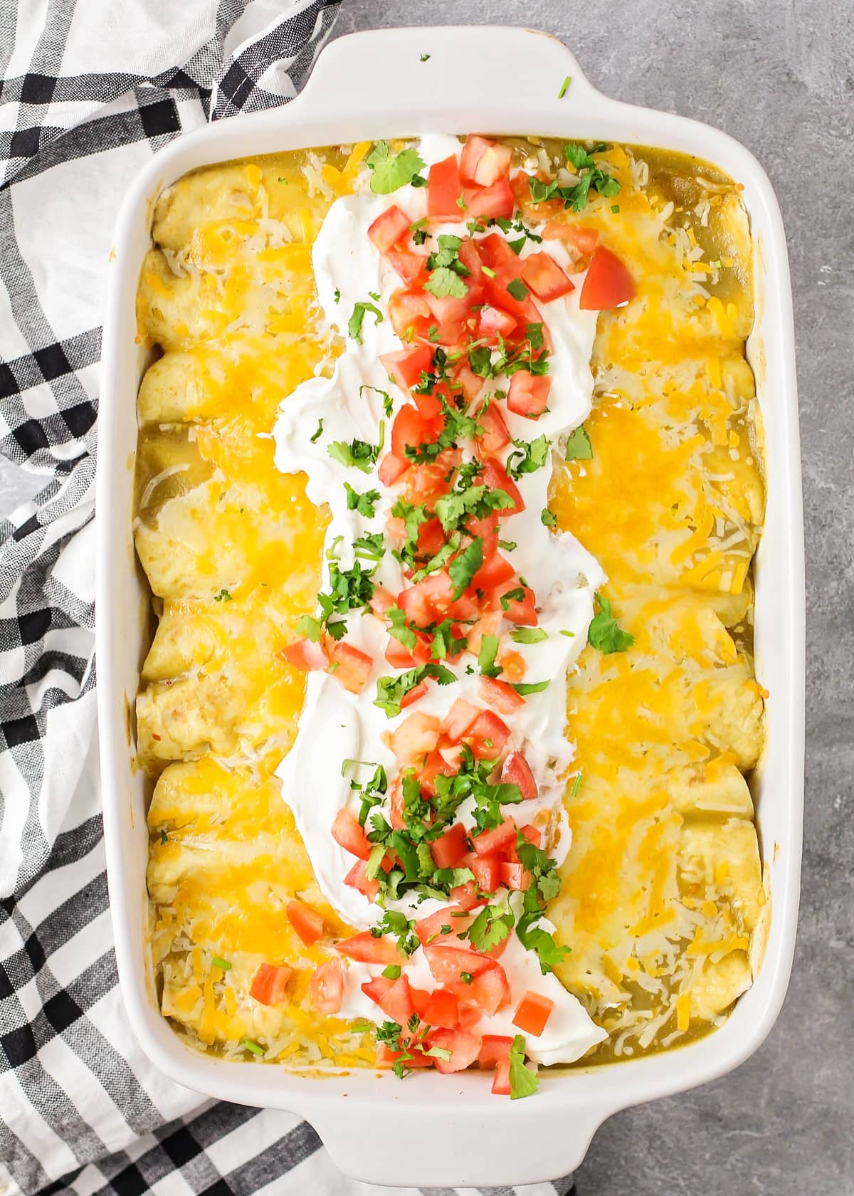 A white baking dish filled with cream cheese chicken enchiladas topped with sour cream, tomatoes, and cilantro.