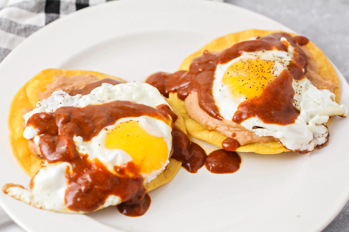 Two corn tortillas topped with refried beans, fried eggs, and red enchilada sauce.