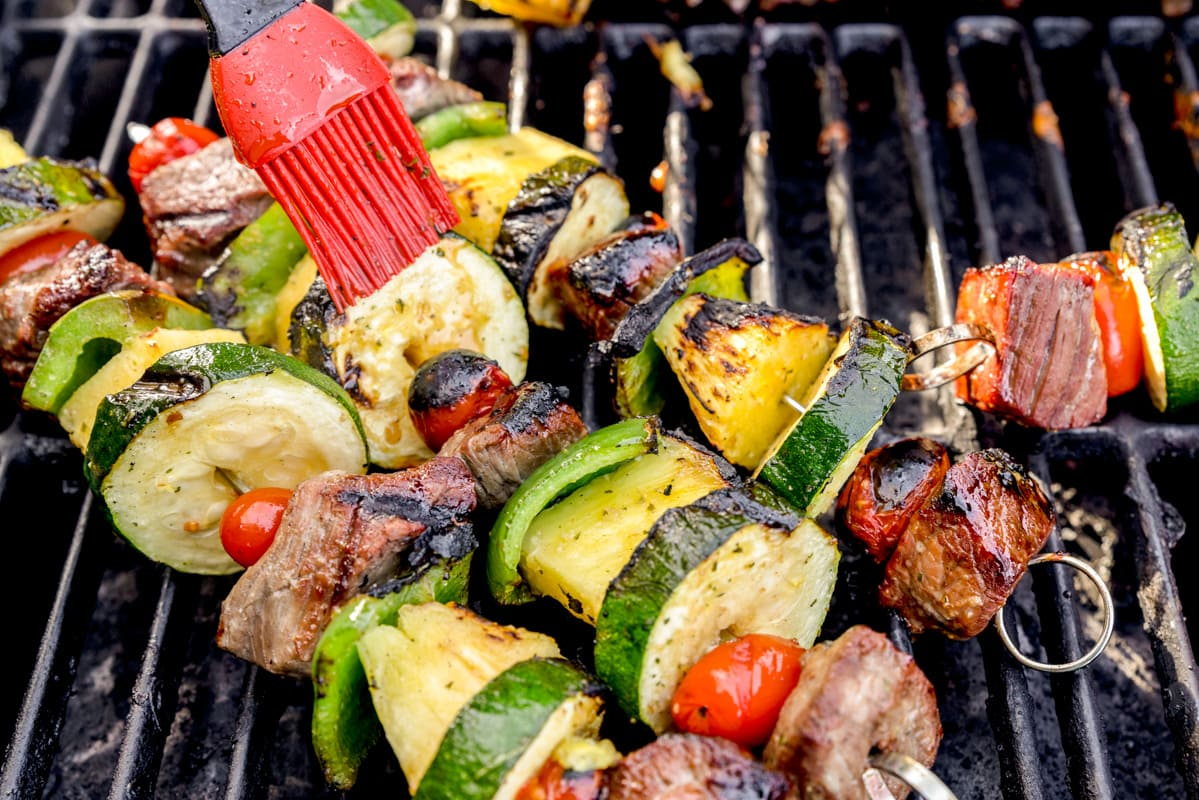 Three vegetable and meat kabobs on a barbecue grill and a basting brush brushing one of the kabobs.