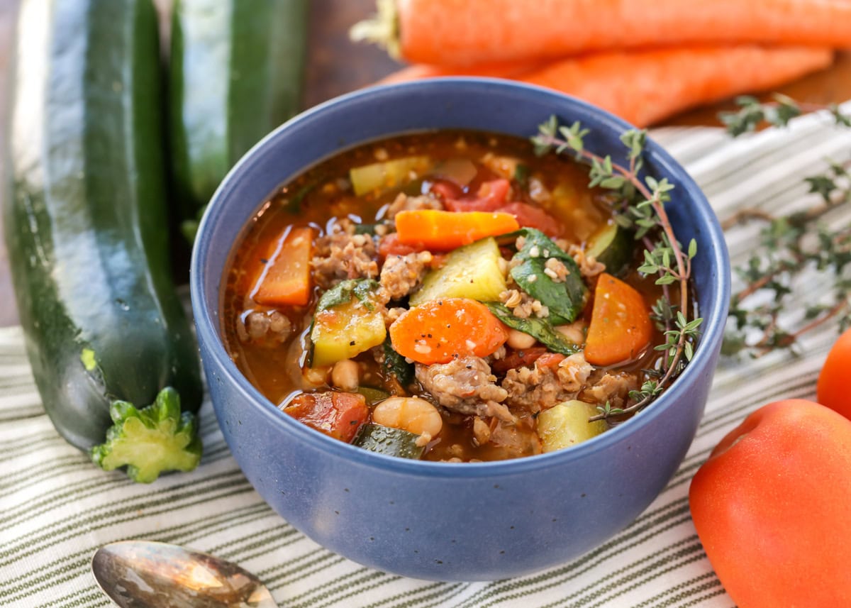 Sausage soup recipe served in blue bowl with veggies all around.