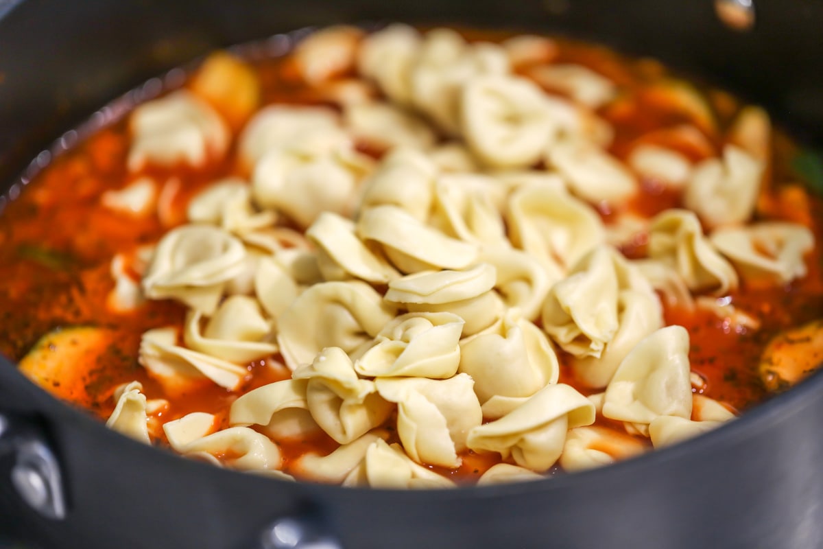 Tortellini being poured into pot with veggies and broth.