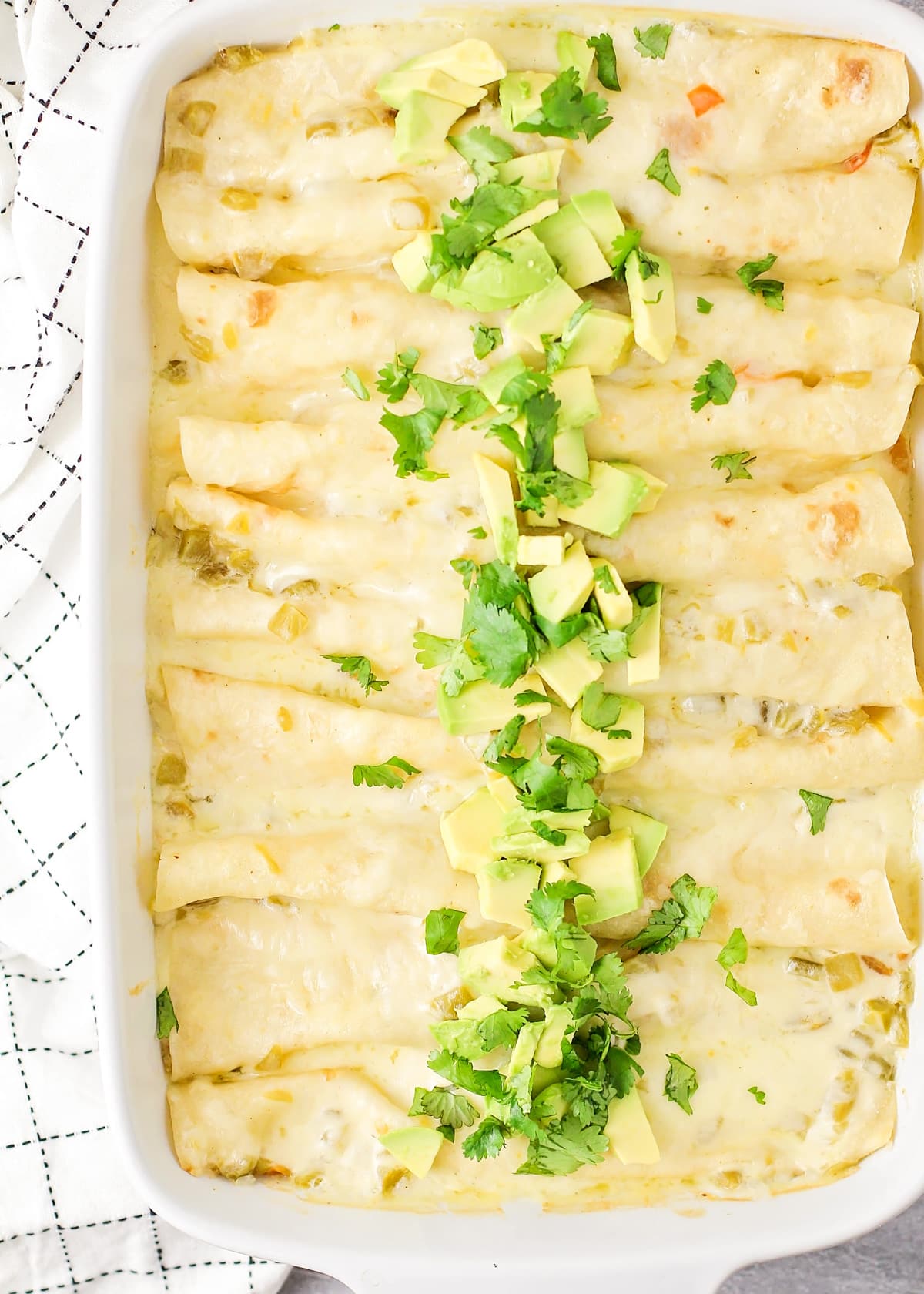 A close up of a baking dish filled with white chicken enchiladas topped with avocado and cilantro.