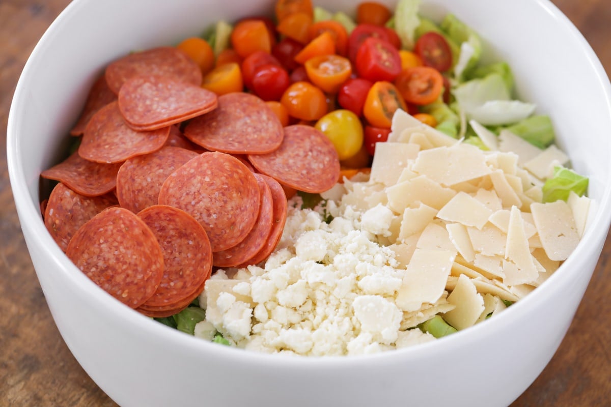 Ingredients for pizza salad in white bowl including cheese, tomatoes, pepperonis and salad.