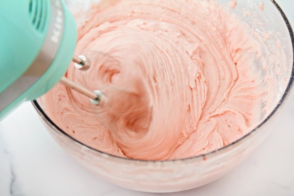 How to make strawberry frosting process pictures - ingredients being mixed with an electric mixer in a clear glass bowl. 