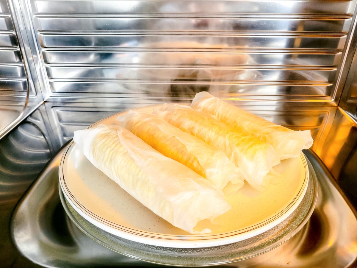 Wrapped cobs of corn on a white plate in the microwave.