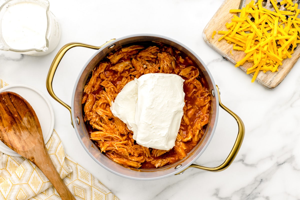 Adding cream cheese to a pot of seasoned shredded chicken in a pot.