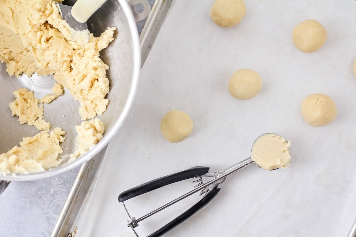 Scooping and dropping sugar cookie dough onto a lined baking sheet.