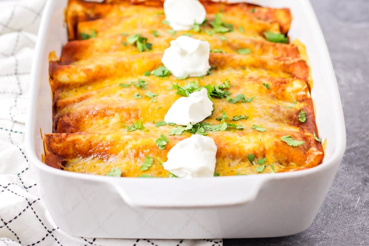 A white baking dish filled with ground beef enchiladas and topped with sour cream.