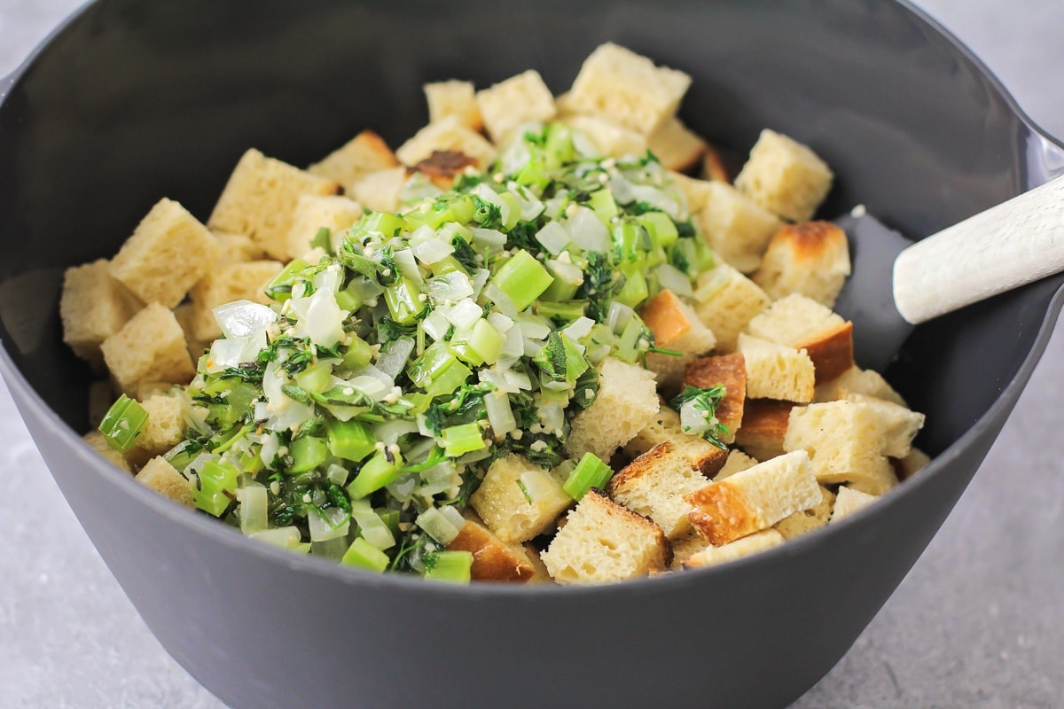 Adding cooked veggies to bread cubes in a grey bowl.