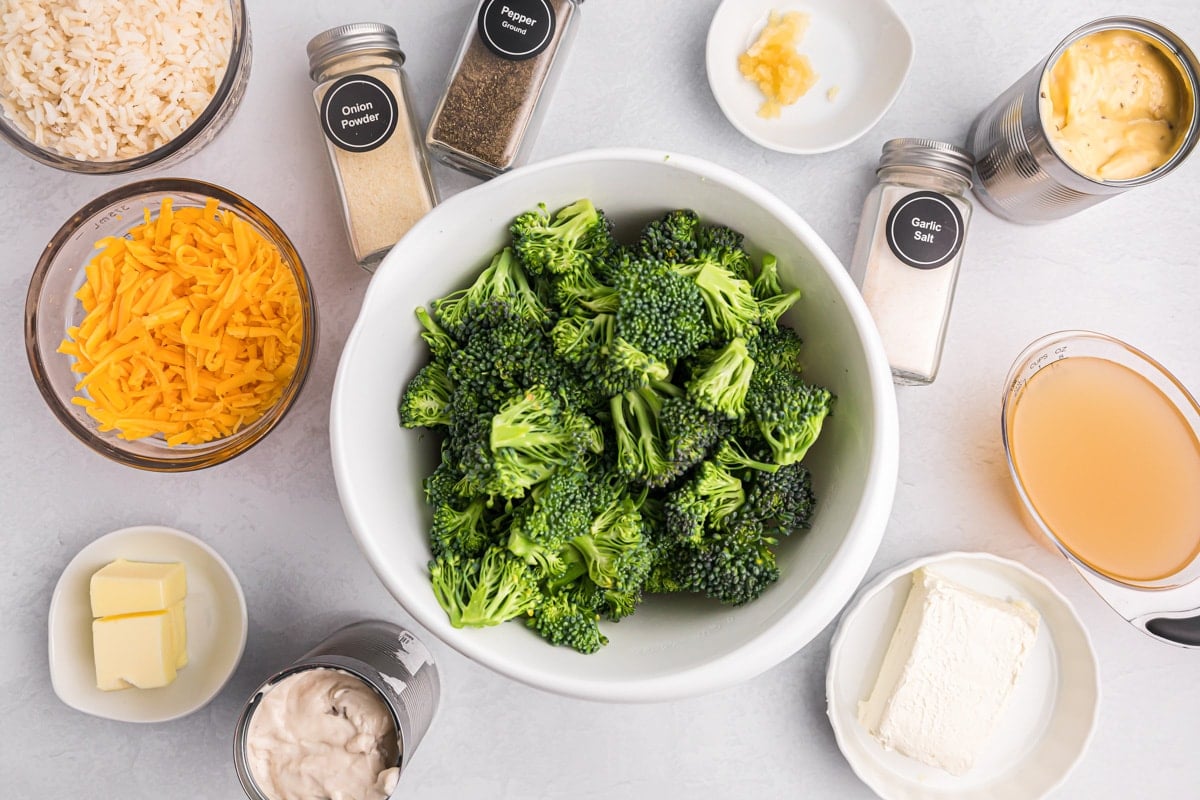 A bowl of broccoli, cheese, cans of soup, and other seasoning ingredients measured on a kitchen counter.
