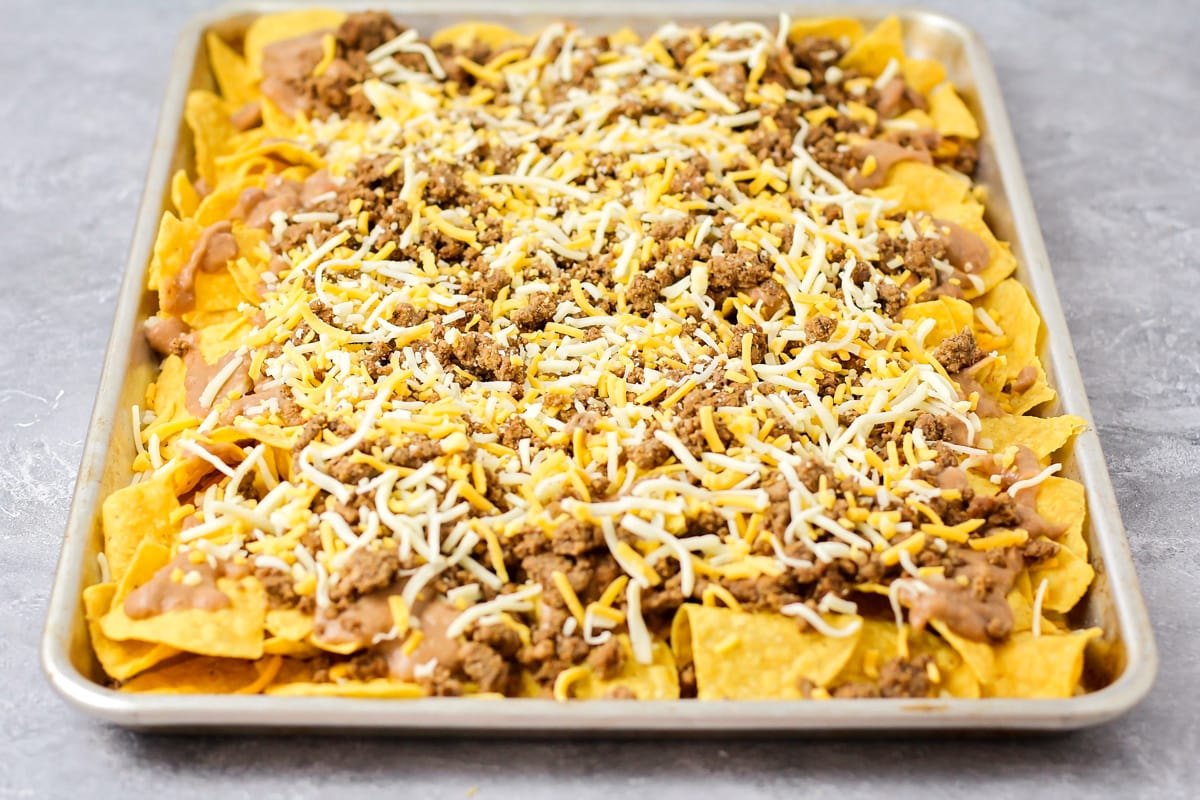 A sheet pan of tortilla chips topped with beans, meat, and shredded cheese.