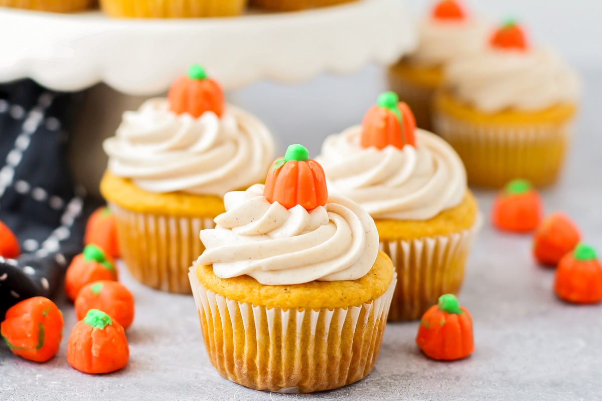 Pumpkin cupcakes topped with cinnamon cream cheese frosting and candy pumpkins on a wire rack.