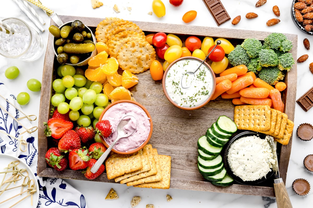 Adding a variety of crackers to a board filled with produce and dips.