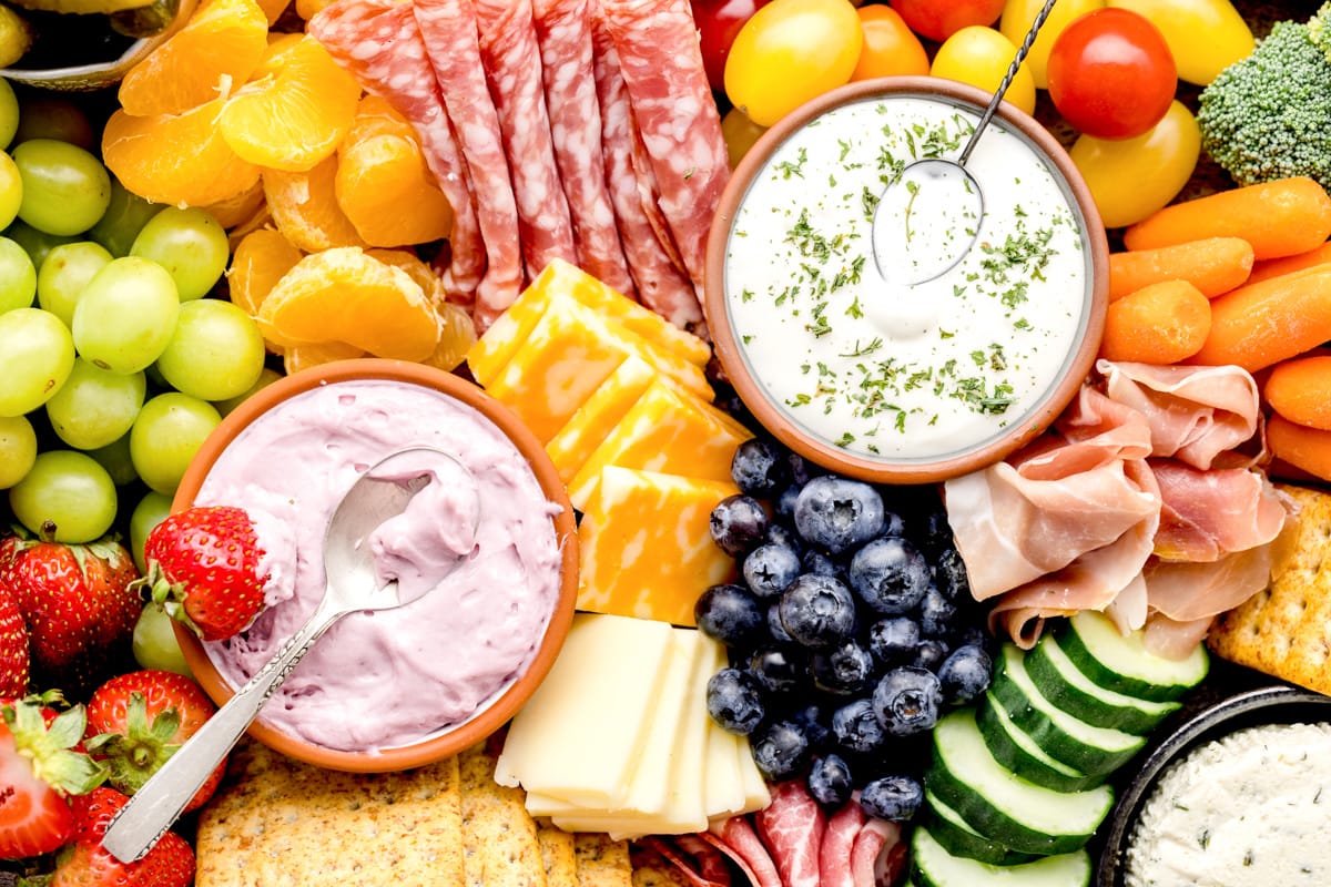 Close up of dips, meat, cheese, and produce on a wooden board.