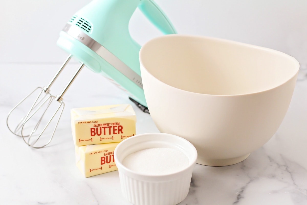 Two sticks of butter next to a bowl of sugar and a mixer.