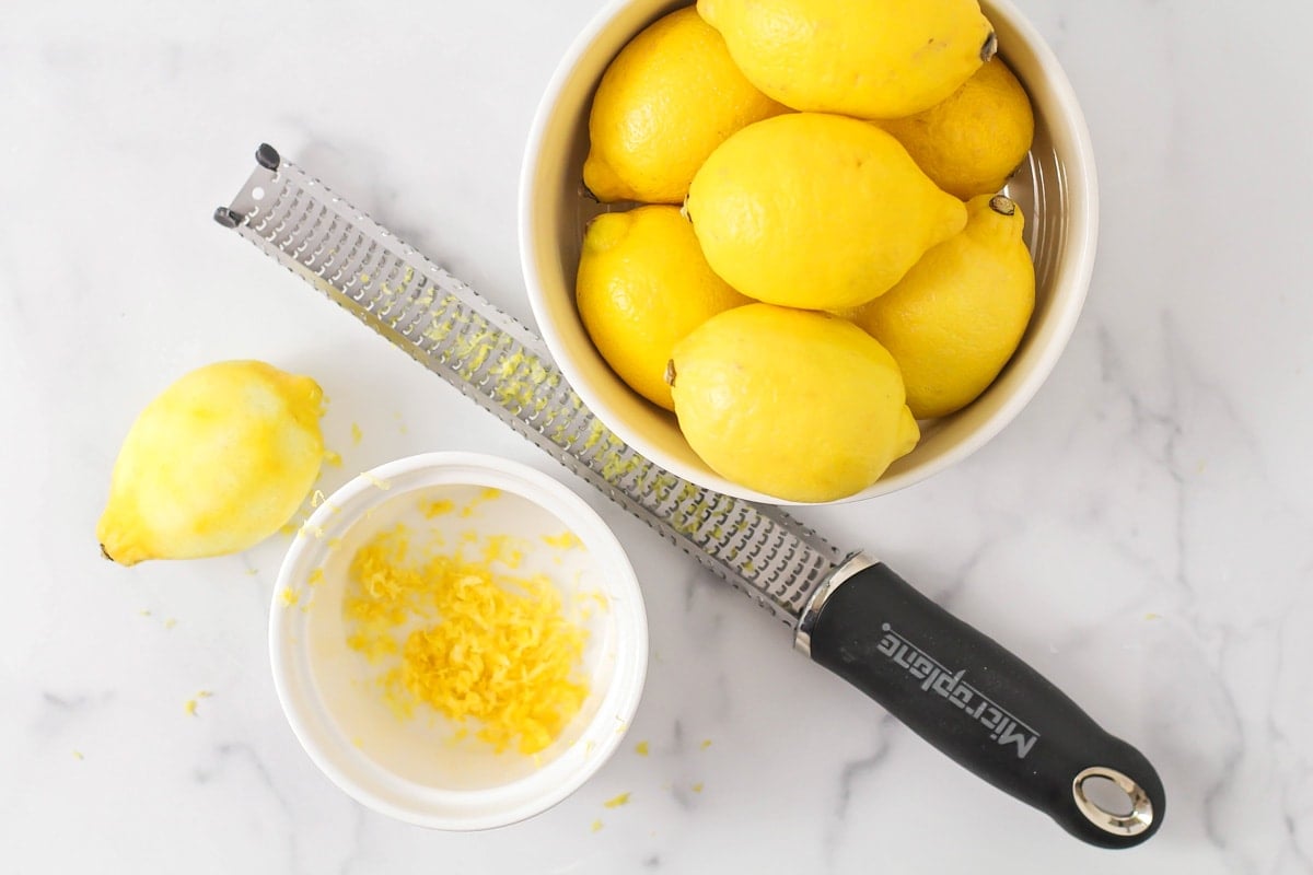 A zested lemon next to a white bowl next to a bowl of lemons.