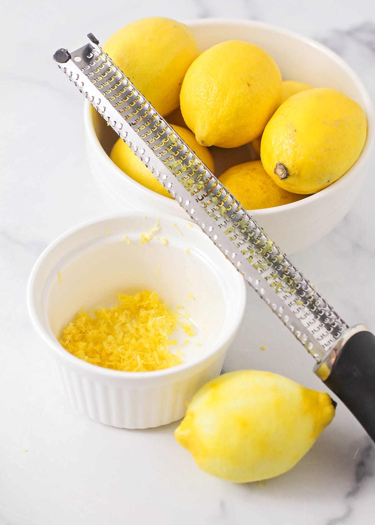 Close up of lemon zest in a white bowl next to fresh lemons.