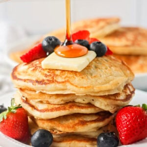 Buttermilk Pancakes recipe stacked on white plate with fruit, butter and syrup on top.
