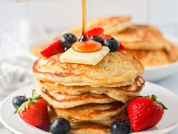 Buttermilk Pancakes recipe stacked on white plate with fruit, butter and syrup on top.