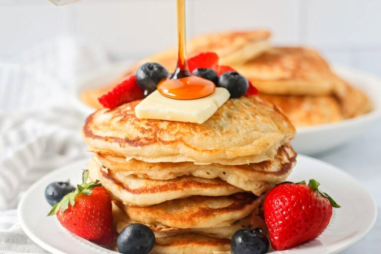 Buttermilk Pancakes recipe stacked on white plate with fruit, butter and syrup on top.