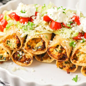 Chicken taquitos stacked in white platter topped with lettuce, tomato and sour cream.