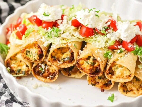 Chicken taquitos stacked in white platter topped with lettuce, tomato and sour cream.