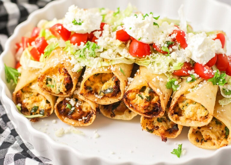 Chicken taquitos stacked in white platter topped with lettuce, tomato and sour cream.