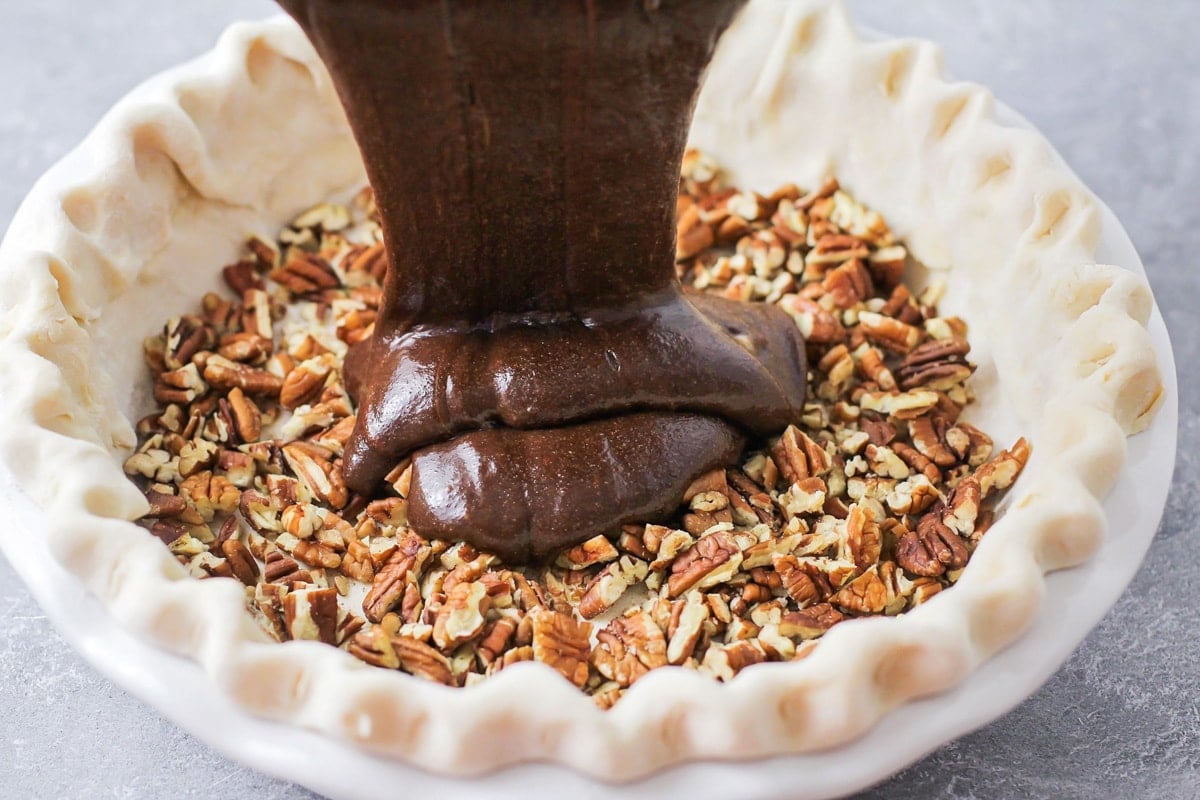Pouring chocolate filling on top of crumbled pecans in a pie crust.