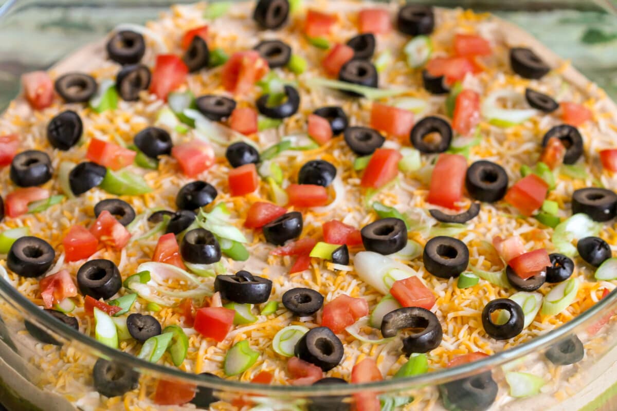 Close up of dip topped with cheese, green onion, olives, and tomatoes.