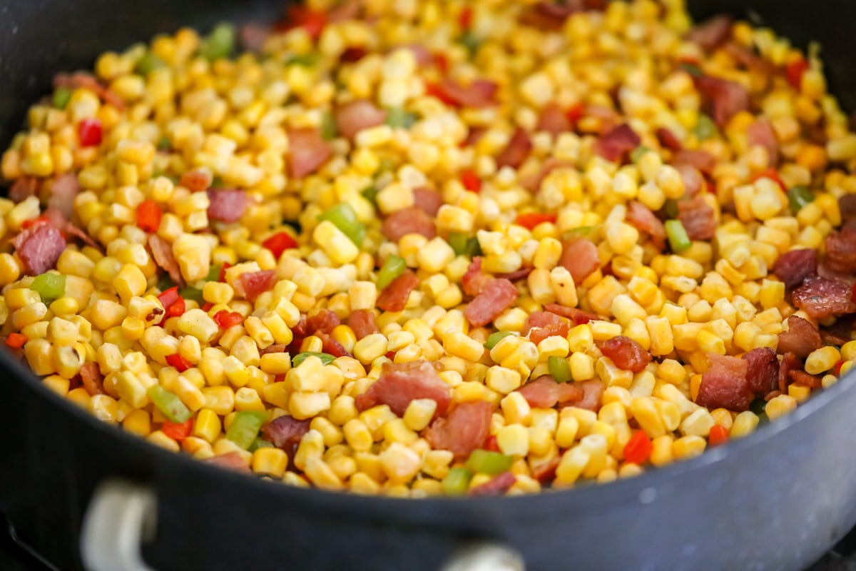 Corn mixed with bacon and peppers in a pan on the stove.