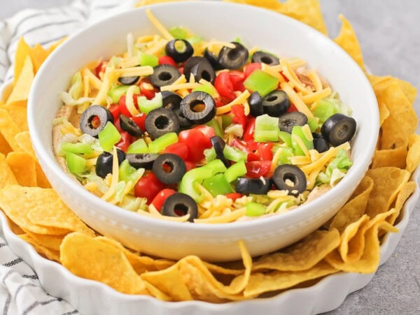 A bowl of taco dip topped with green olives, tomatoes, and lettuce, served with tortilla chips.