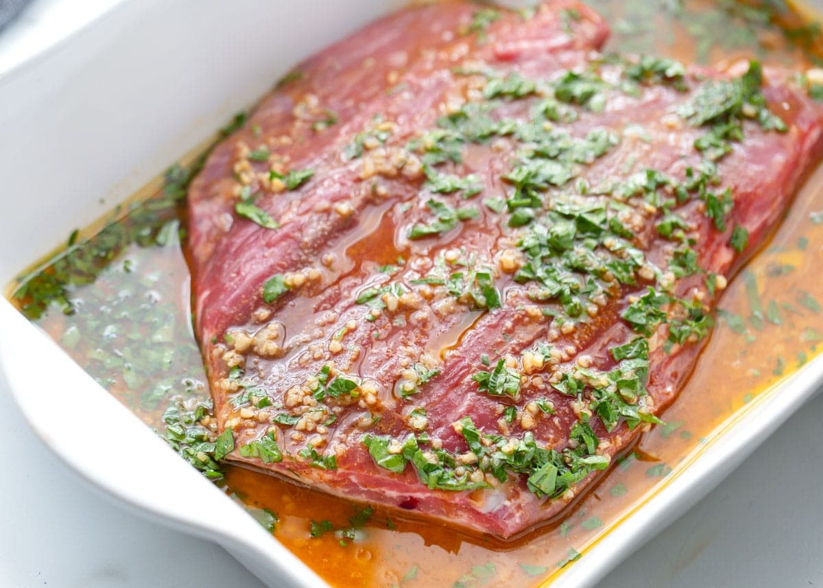 A flank steak marinating in a white dish.
