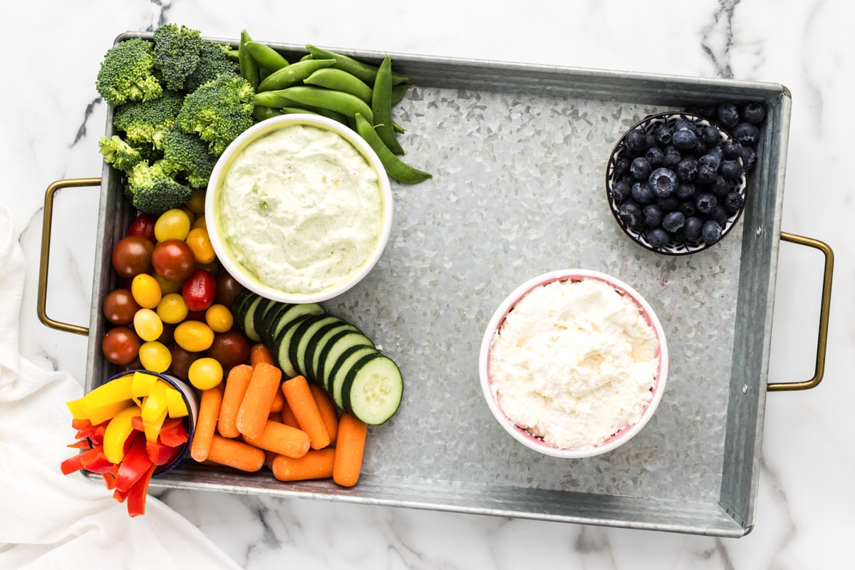 Adding veggies to a metal tray topped with dips.