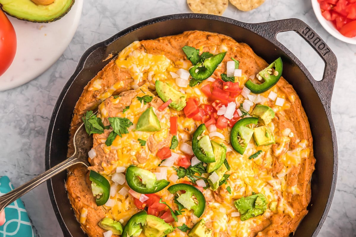 Bean dip in a skillet topped with shredded cheese, tomatoes, onions, and jalapenos.