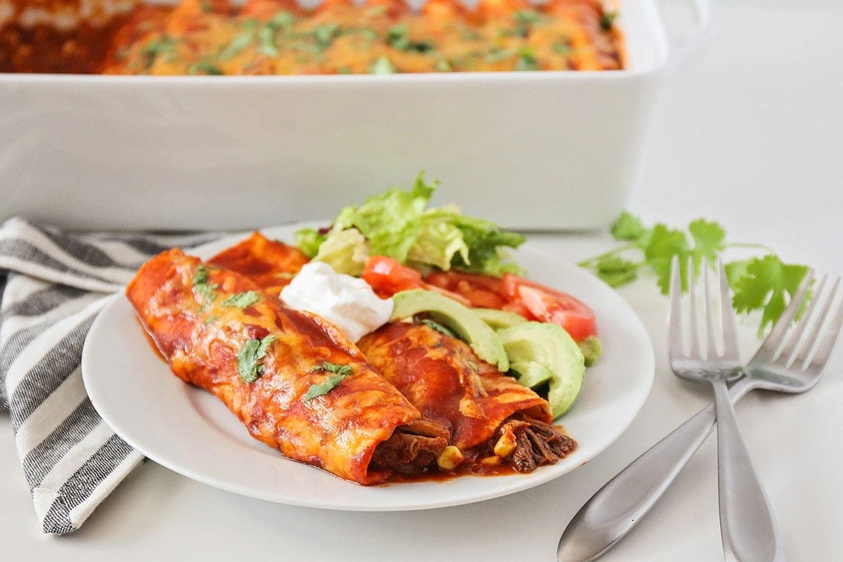 Beef Enchiladas topped with sour cream and avocado slices on a white plate.