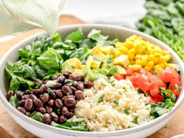 A burrito bowl topped with fresh veggies with a dressing being added.