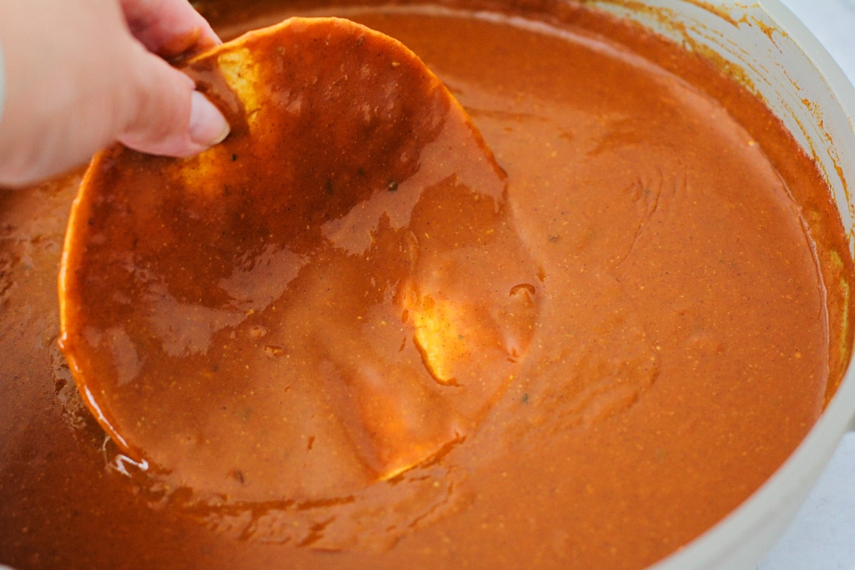 Corn tortilla being dipped in enchilada sauce.