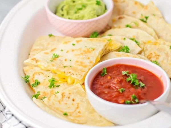 A cheese quesadilla sliced into triangles and served with cups of red and green salsa.